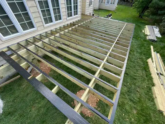 A construction worker framing a wooden deck attached to a house, with tools and materials scattered around.